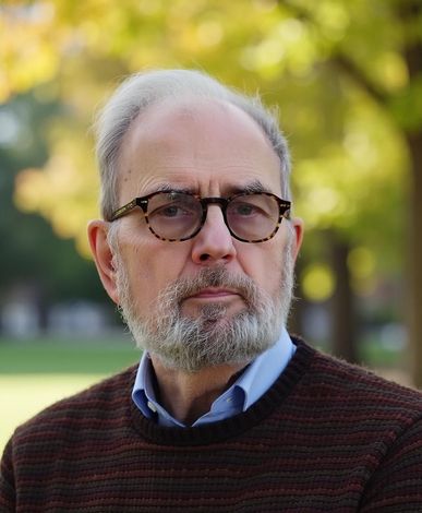 Man with glasses and a beard, in a brown sweater and a blue shirt, outside with a blurred green background.
