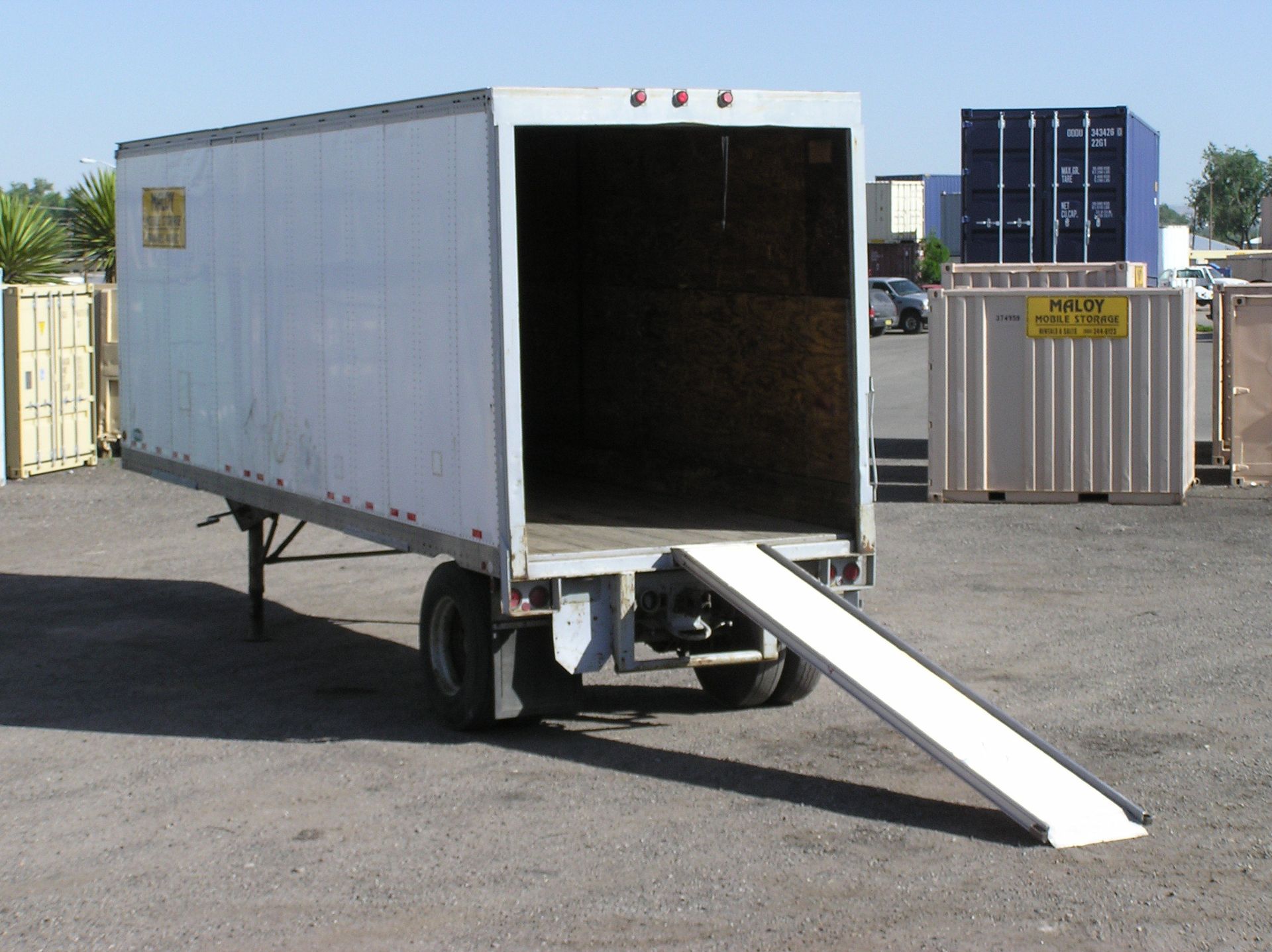 White truck trailer with ramp extended, open in a gravel lot.