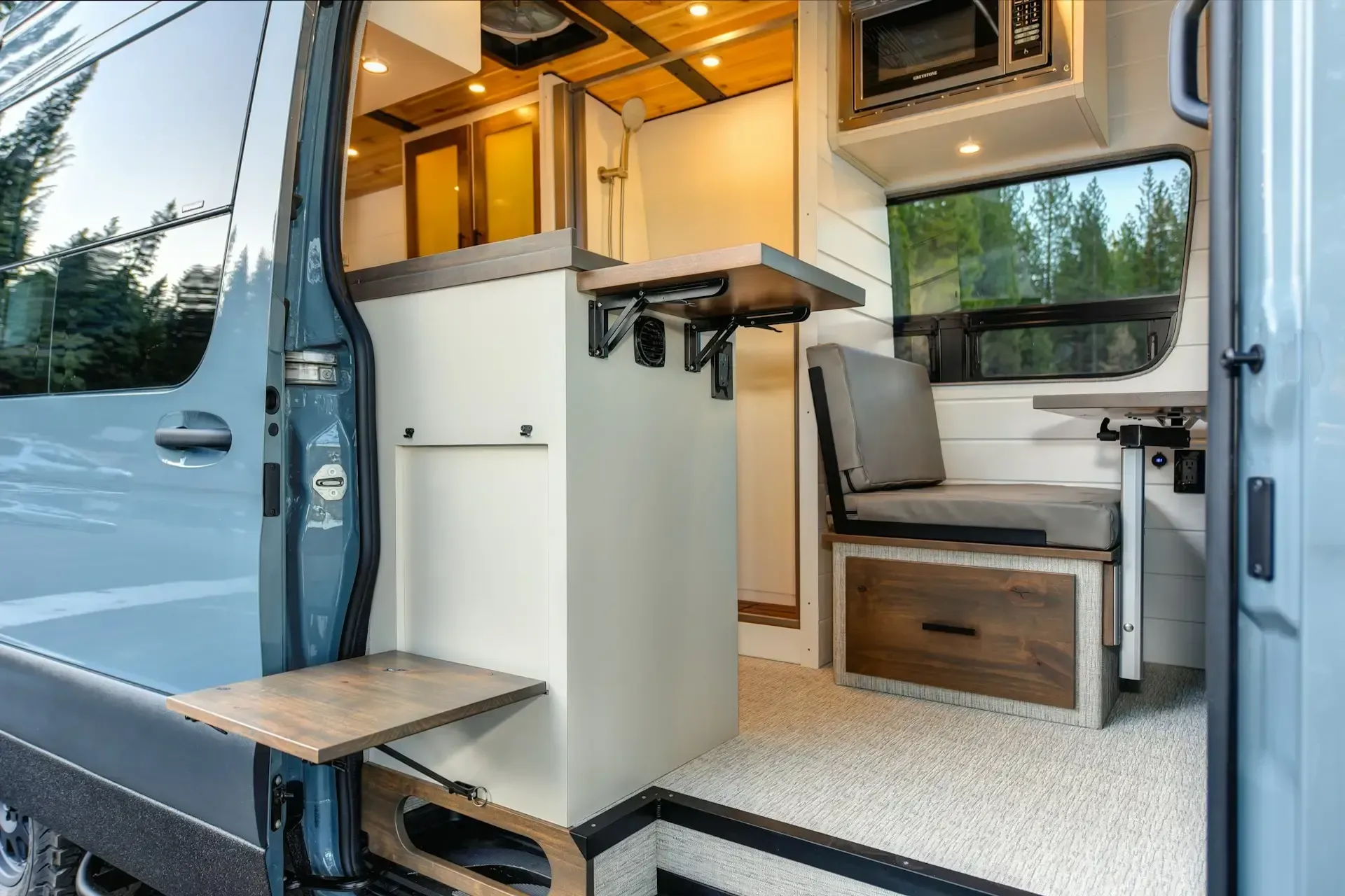 Interior of a camper van with a kitchen area, seating, and a fold-down table. Neutral colors with wood accents.
