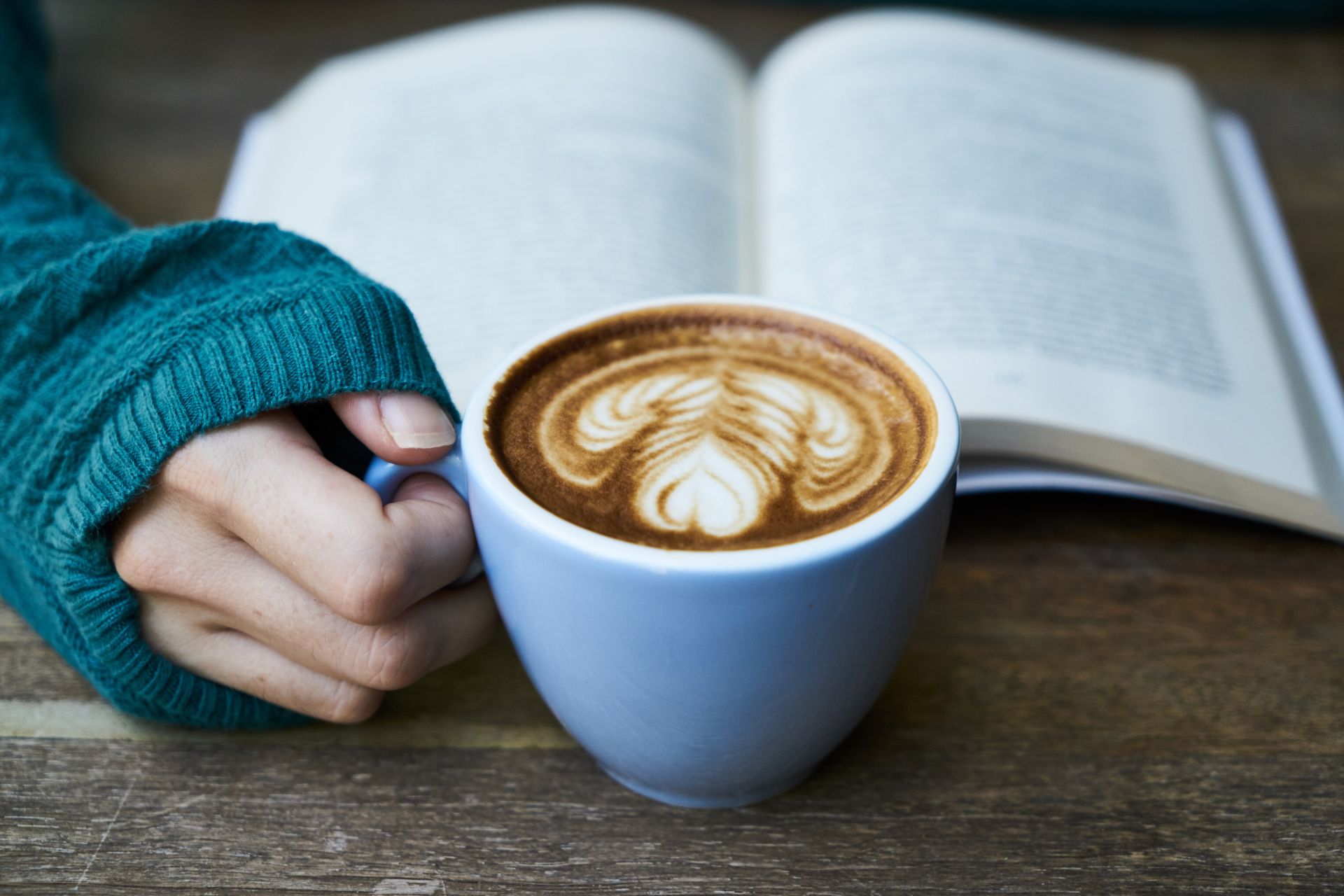 Hand holding a light blue mug of coffee with latte art, next to an open book, on a wooden table.