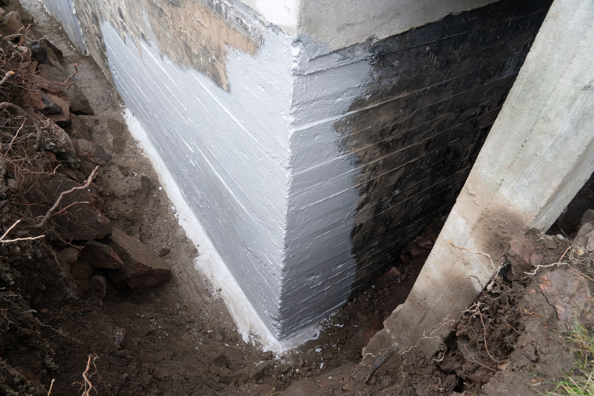 An excavated corner of a concrete foundation with a patch of gray waterproof sealant applied to the surface.