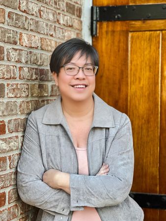A woman is leaning against a brick wall with her arms crossed and smiling.