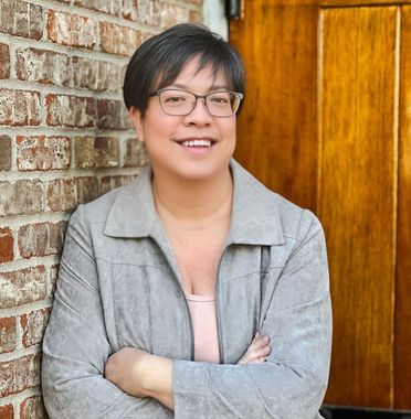 A woman wearing glasses is leaning against a brick wall with her arms crossed.