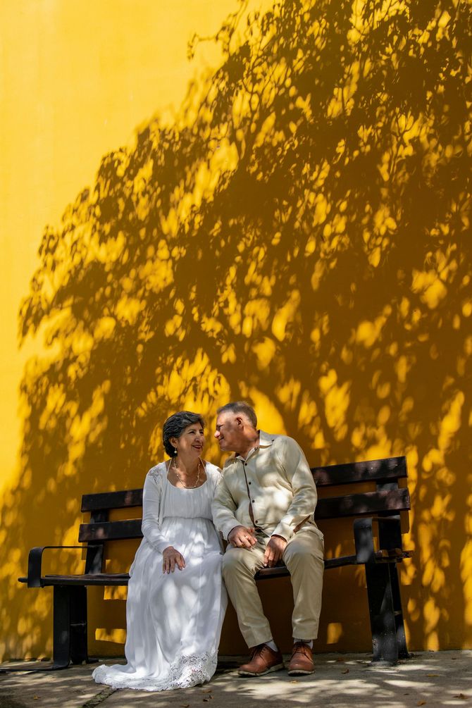 A bride and groom are sitting on a bench in front of a yellow wall.