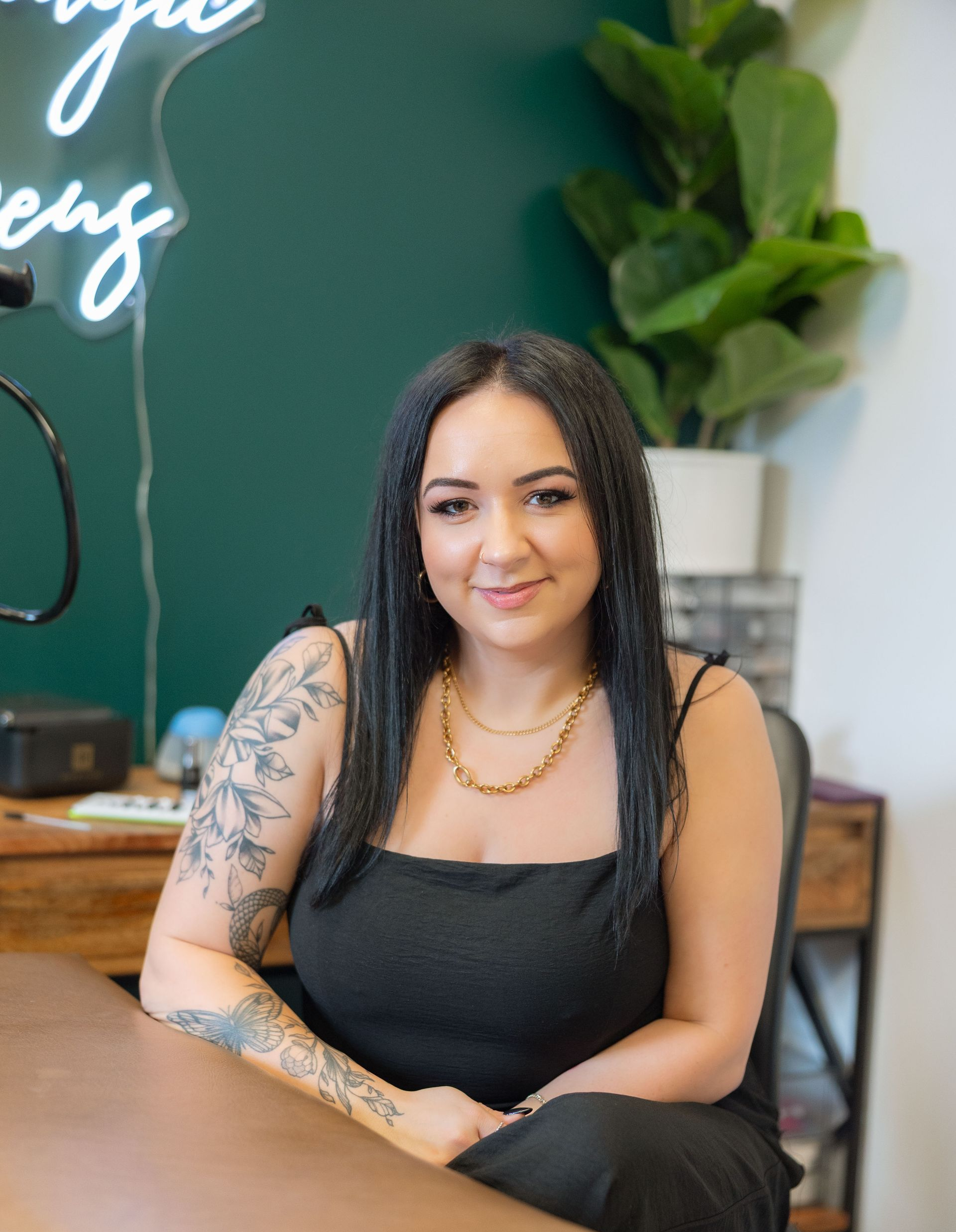 Woman with arm tattoo, gold necklace, and black top sitting in front of a green wall with neon sign.