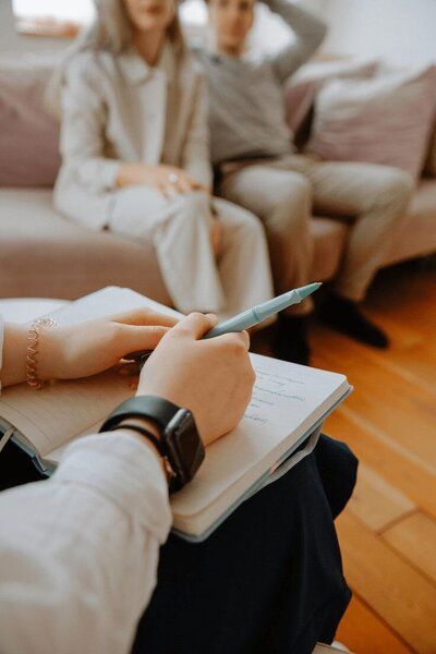 A woman is writing in a notebook while a man and woman sit on a couch.