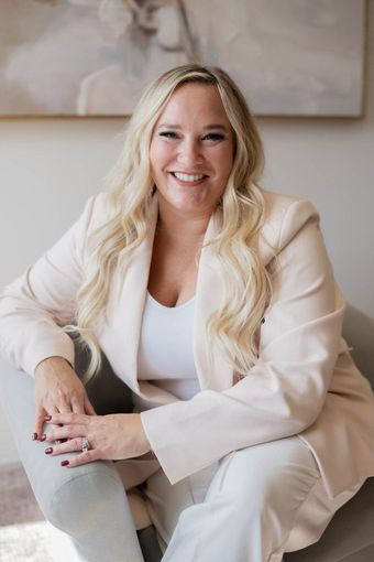 A woman in a white jacket and white pants is sitting in a chair and smiling.