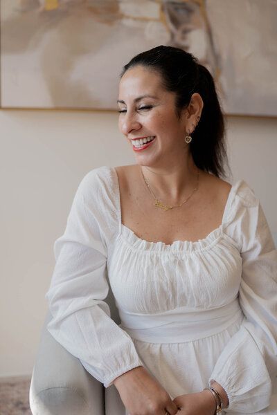 A woman in a white dress is sitting on a couch and smiling.