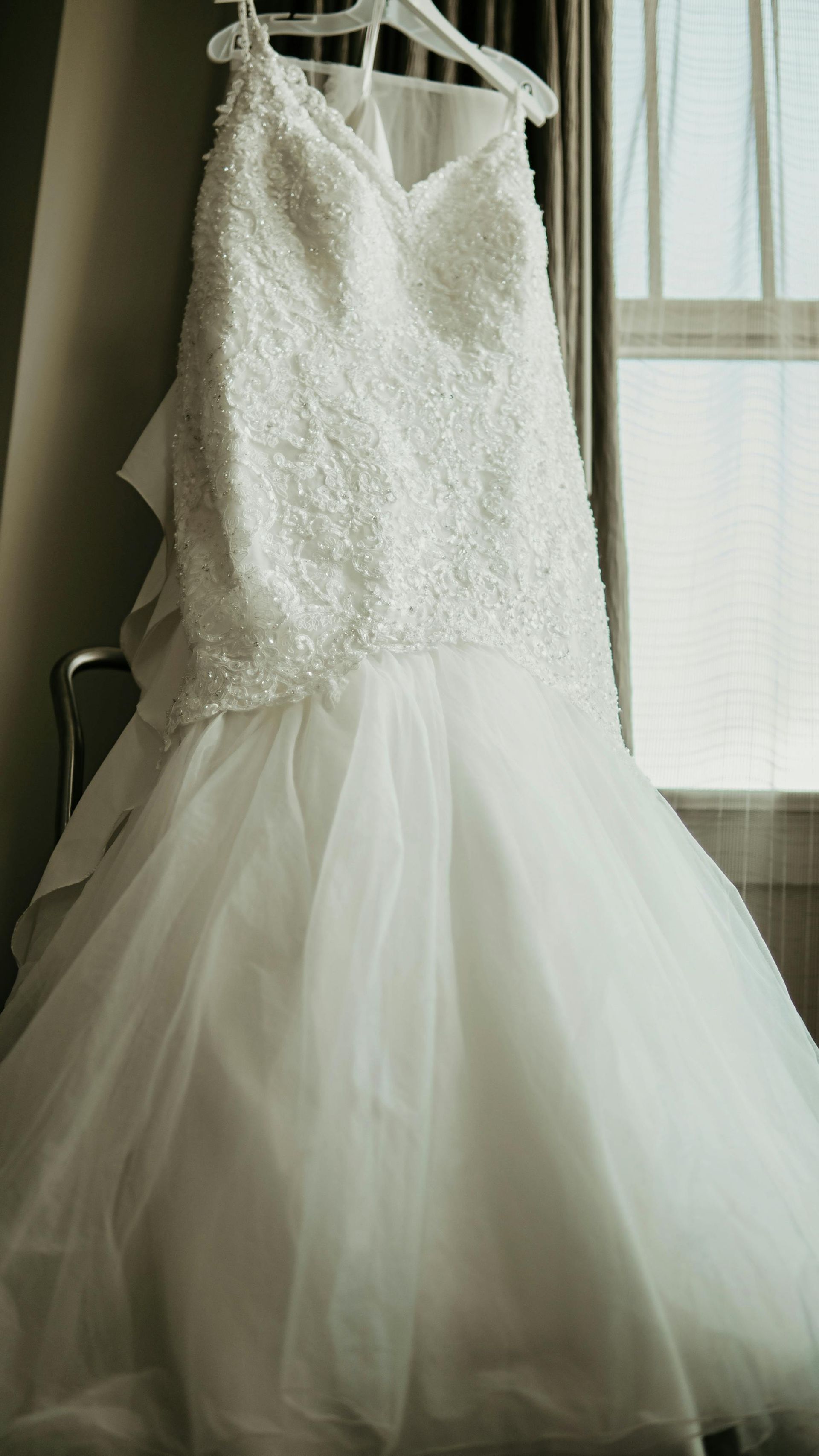 A wedding dress is hanging on a hanger in front of a window.