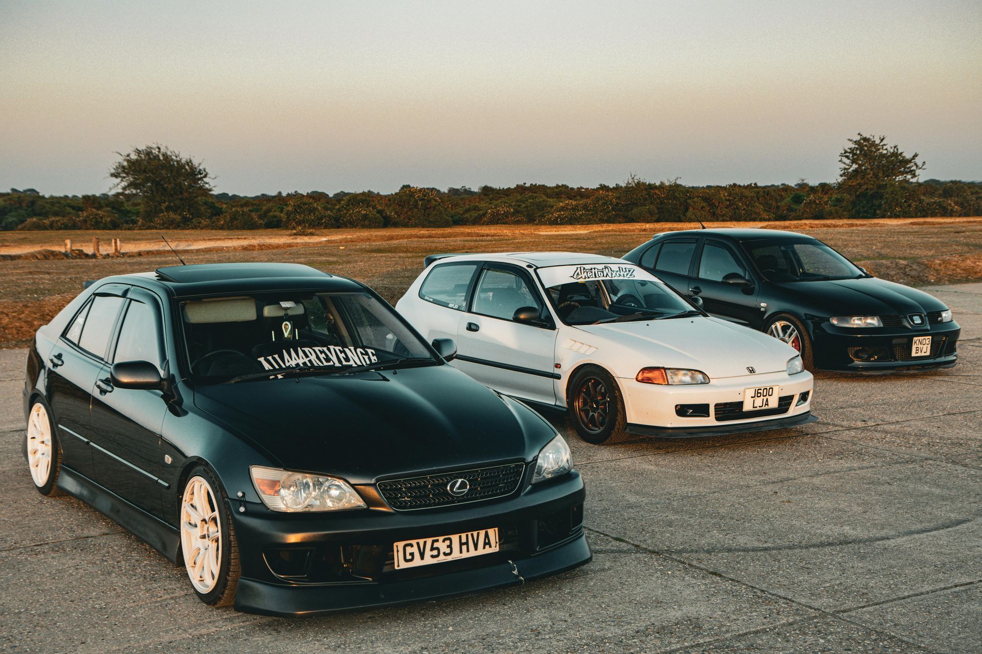 Three modified cars parked on asphalt at sunset: black Lexus, white Honda, and dark gray car.