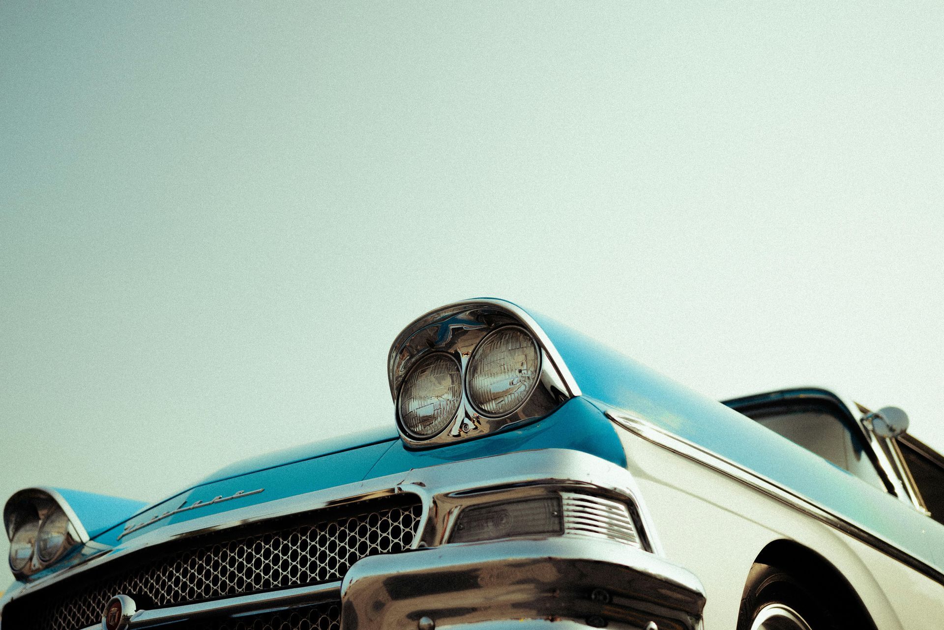 Blue and white classic car, close-up of front. Headlights, grill, and hood visible against a light blue sky.