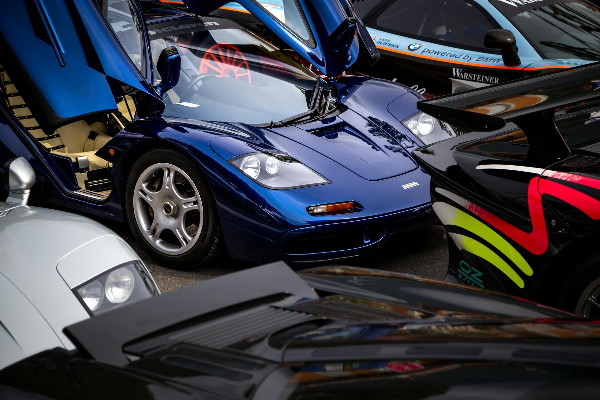 Blue McLaren F1 supercar with open doors, surrounded by other luxury cars, parked.