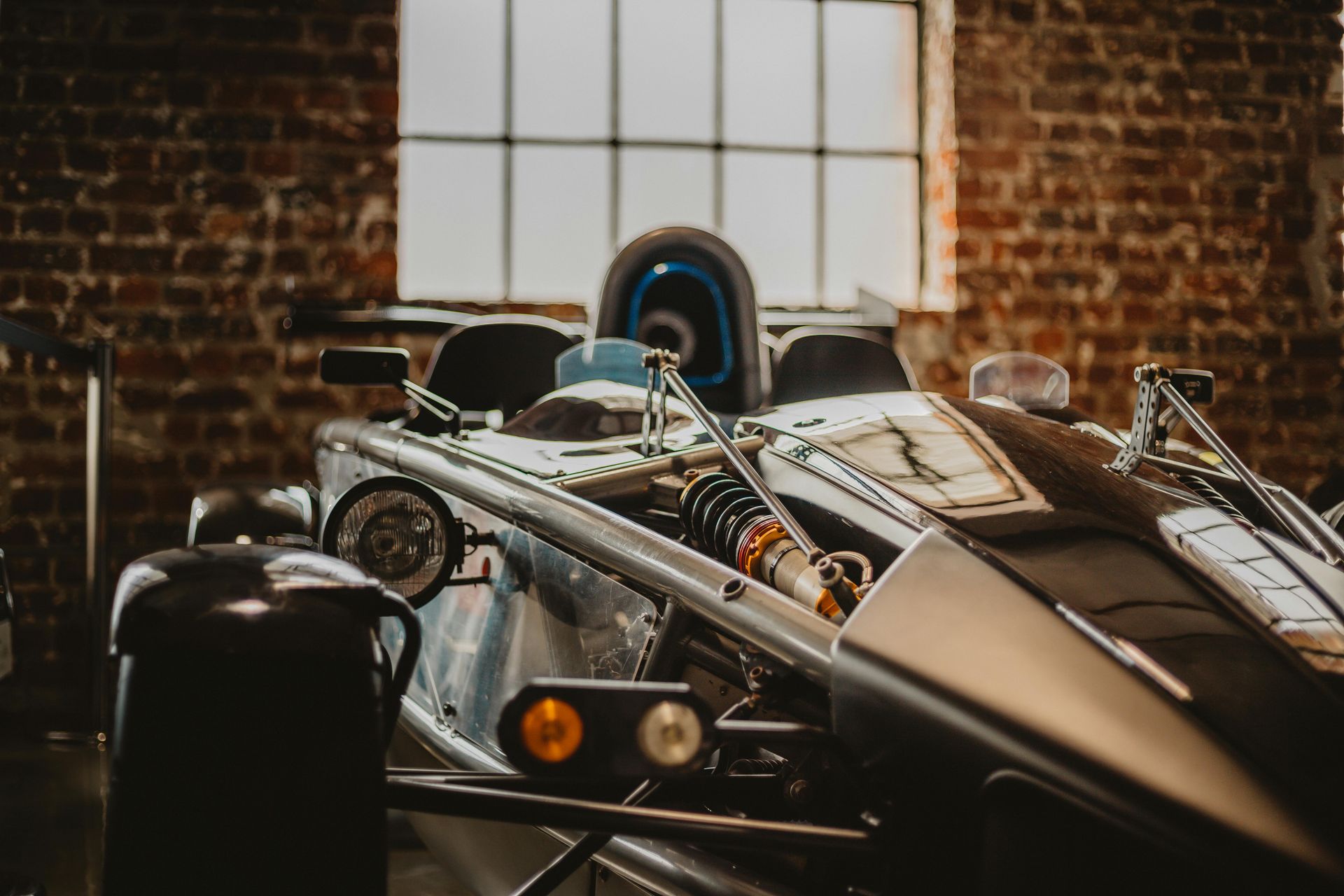 Silver sports car with exposed chassis in a brick-walled building, tire visible in the back.