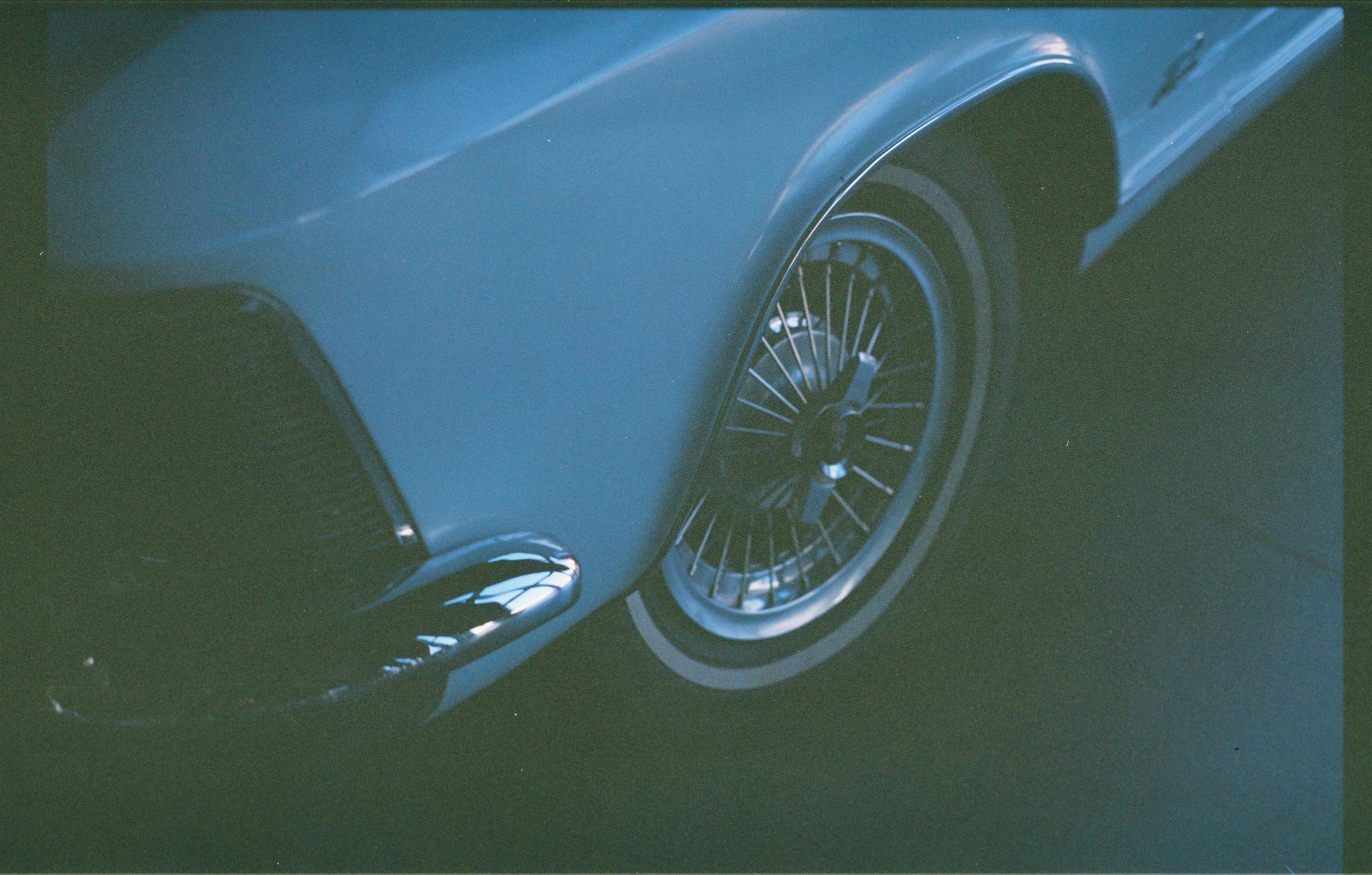 Close-up of a white vintage car's front fender and wire wheel with white wall tire, shadowed in blue light.