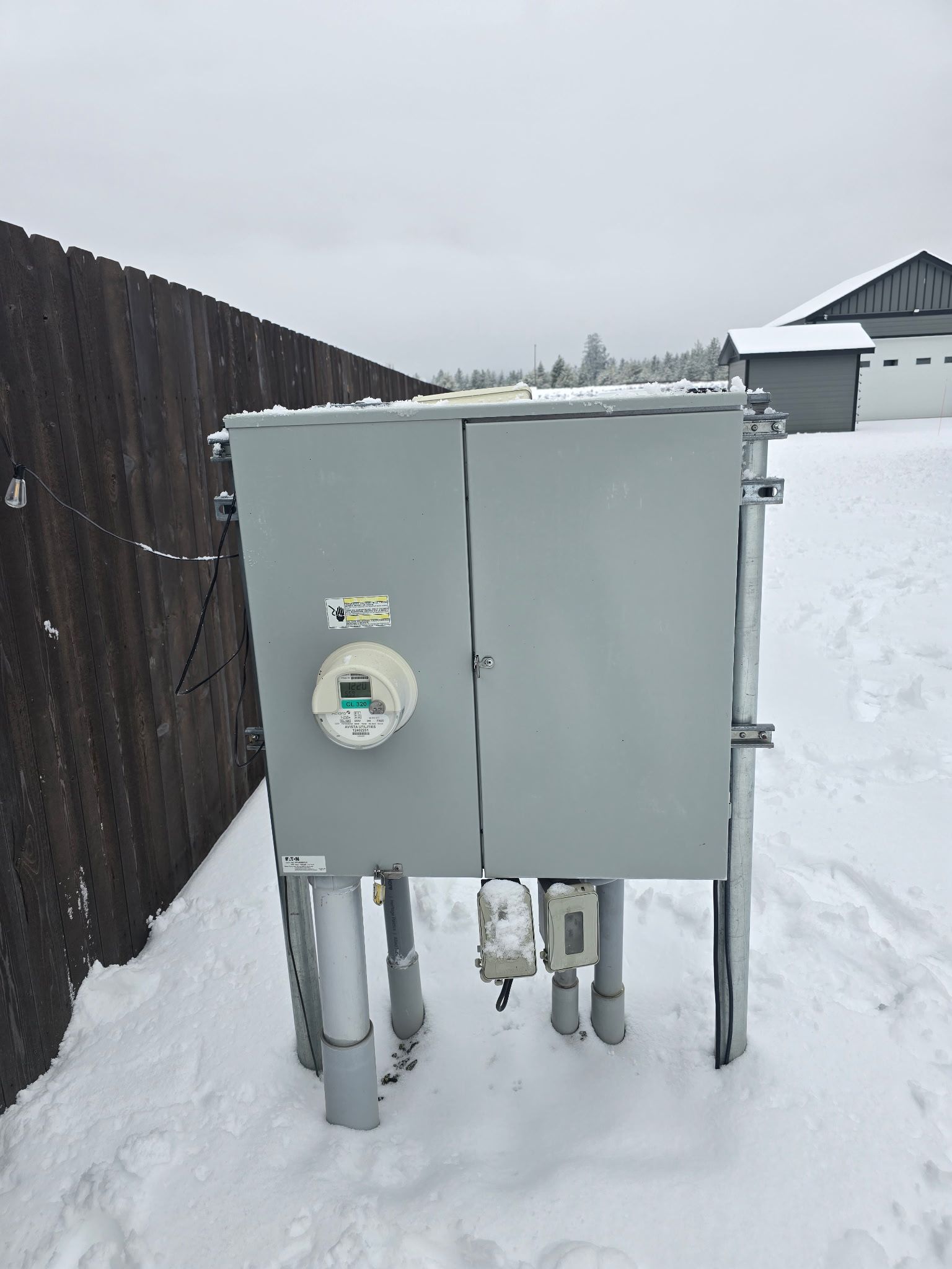 Gray electrical box with a meter, snow-covered ground, next to a brown fence.