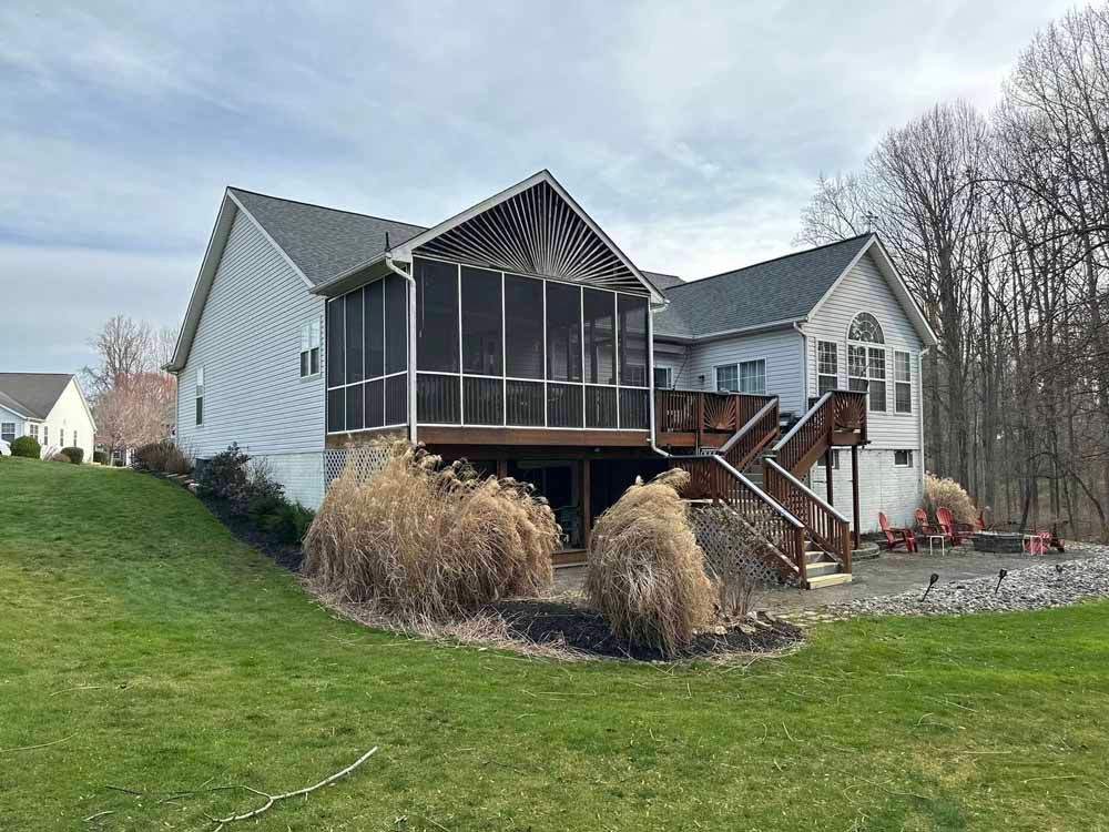 House with screened porch, deck, and sloping yard, trees in background, overcast sky.