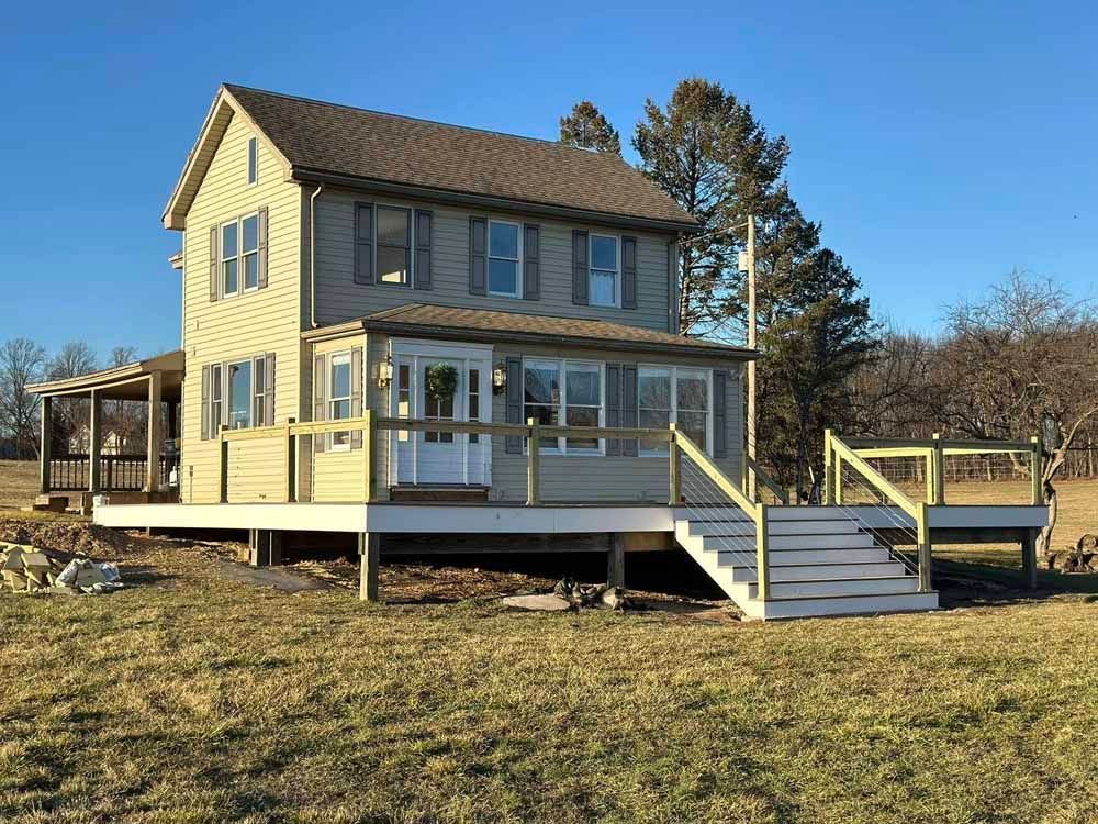 Two-story house with a wooden deck and steps; tan siding, brown roof, and clear blue sky.
