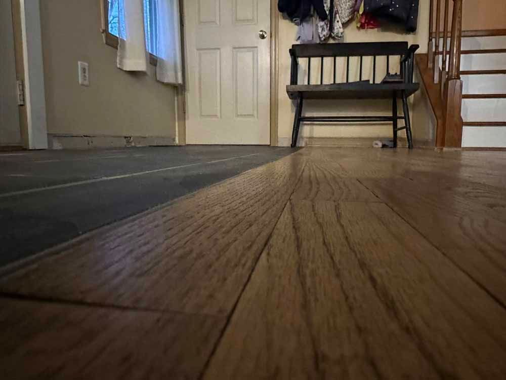 Wooden floor transitions to a dark, unfinished surface. A bench sits against a wall with a door and staircase in the background.