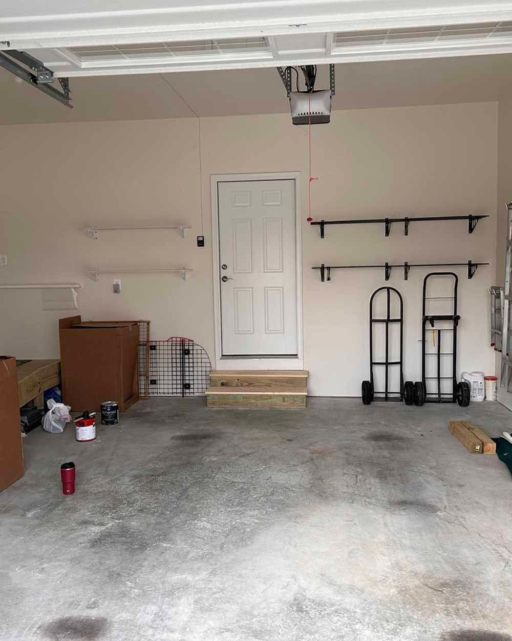 Empty garage interior with white door, concrete floor, shelves, and two hand trucks.