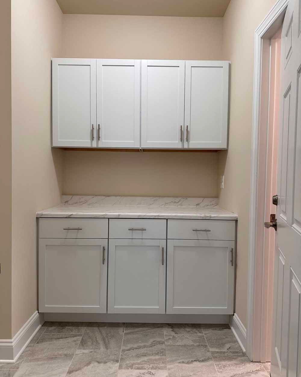 White cabinets and countertops in a utility room. Door to the right. Beige walls and gray tile floor.