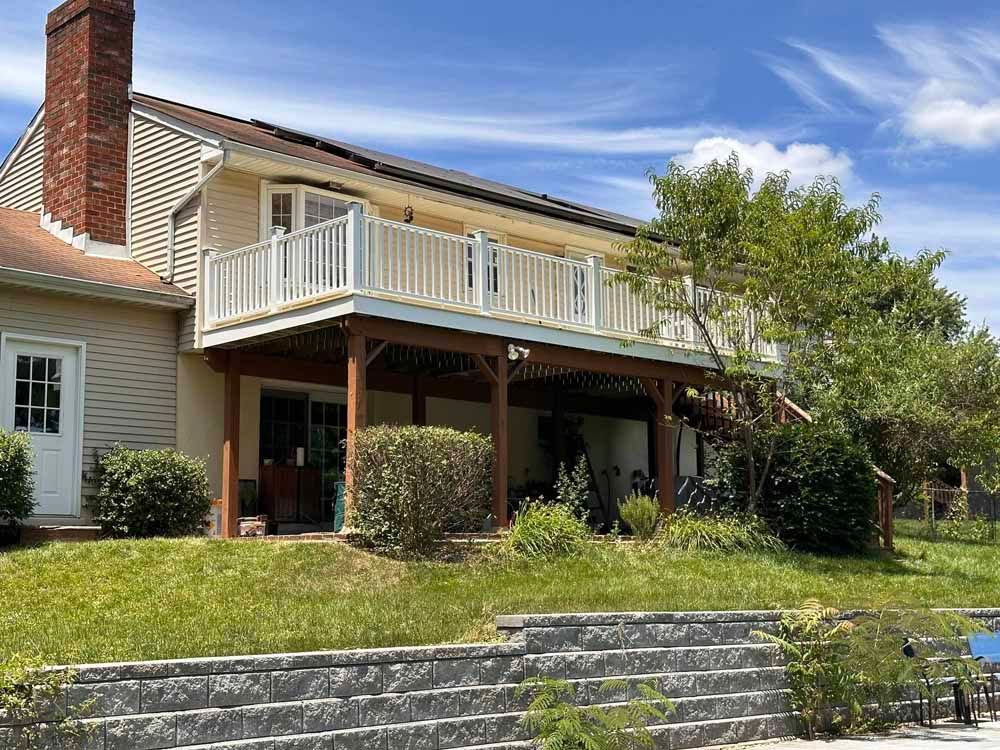 Two-story house with deck, brown siding, white railings, and brick chimney. Green grass and retaining wall in the foreground.