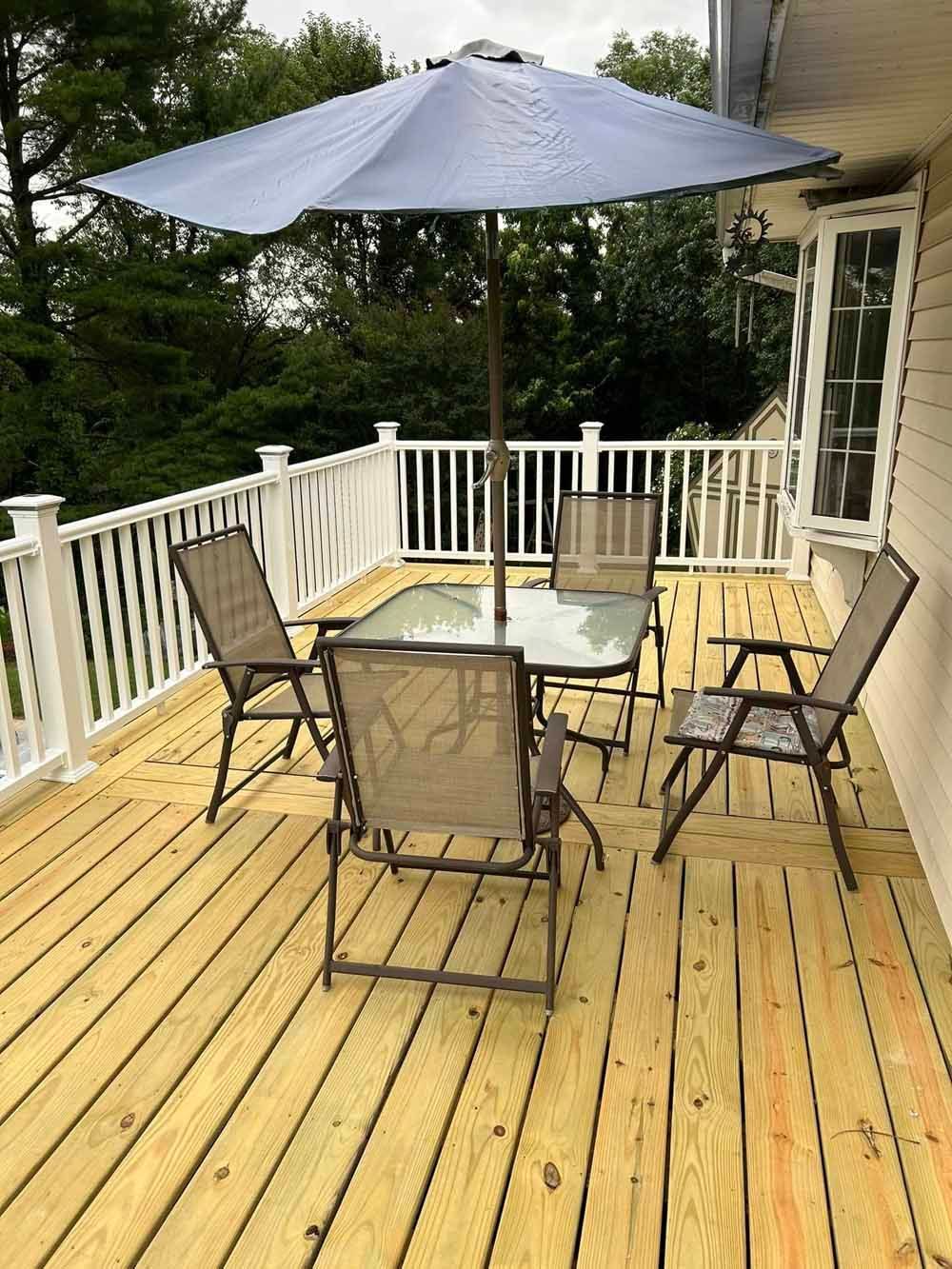 A deck with a table, chairs, and umbrella. White railing. Green trees in the background.