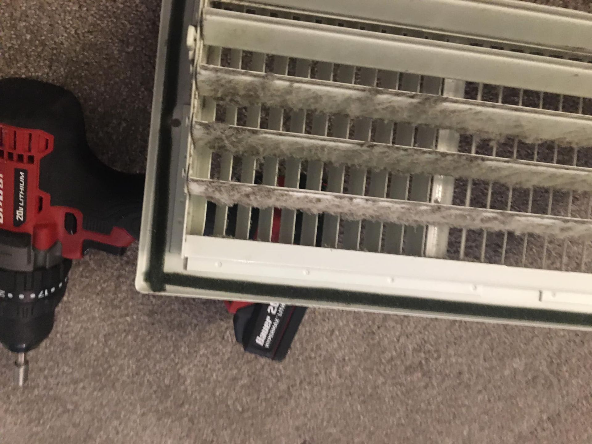 A red drill is sitting next to a dirty air vent.