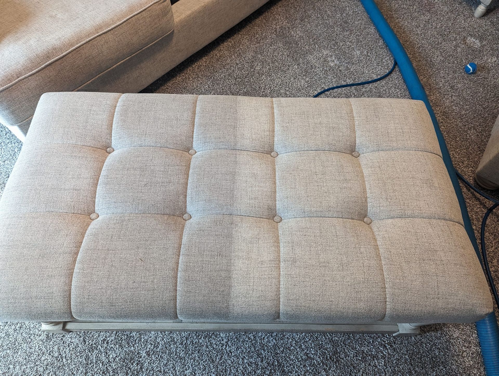 A grey ottoman is sitting on a carpet next to a couch.