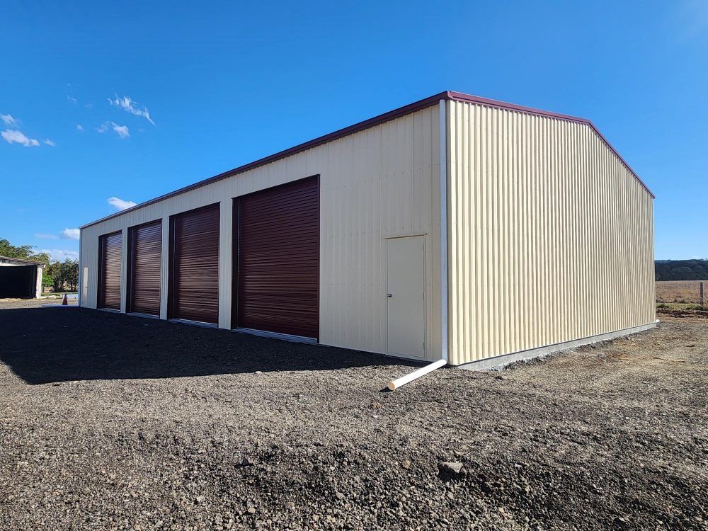A Large Metal Building With Brown Doors Is Sitting In The Middle Of A Dirt Field — Whit Coast Sheds in  Gregory River, QLD
