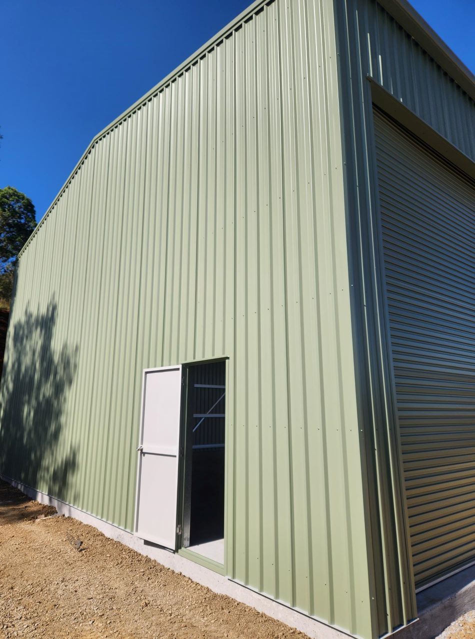 A Green Metal Building With a White Door and Roller Shutters — Whit Coast Sheds in Gregory River, QLD