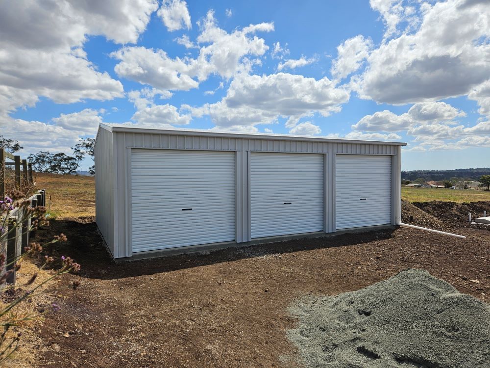 A Garage With Three White Garage Doors Is Sitting On Top Of A Dirt Field — Whit Coast Sheds in Moranbah, QLD