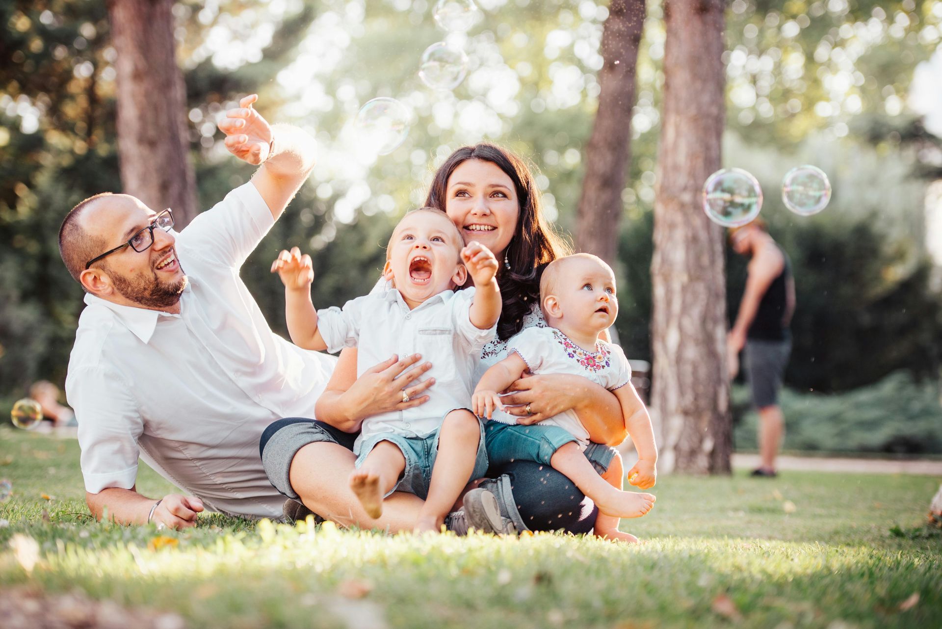 Family in park, bubbles floating. Man points, woman holds two children, all smiling. Green grass, trees.