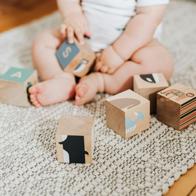 Baby with wooden blocks on a white rug; blocks have letters and animal illustrations.