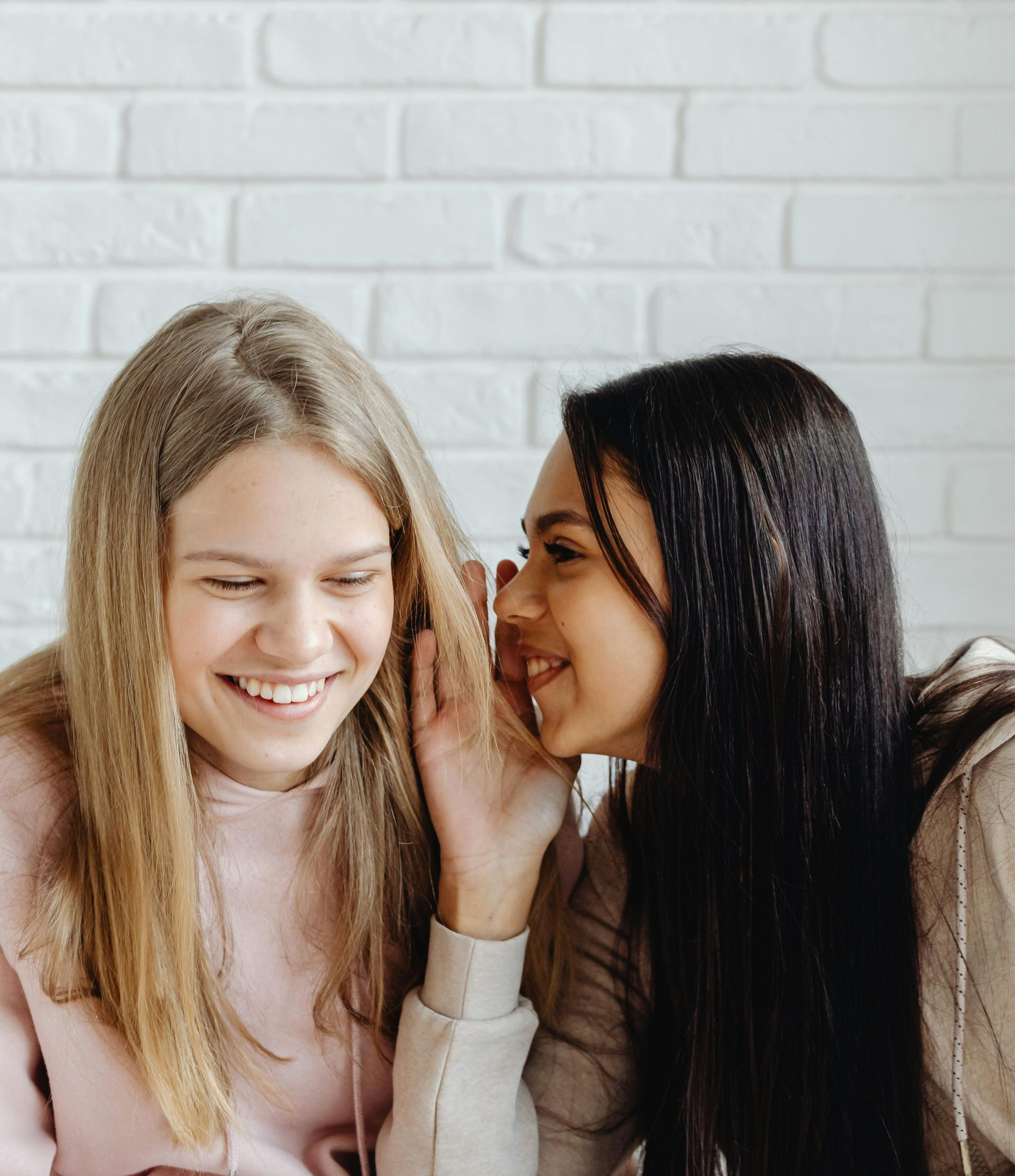 Two women whispering secrets, one laughing, against a white brick wall background.