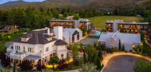 Aerial view of a white mansion with additional buildings, pool, and vineyard nestled amongst trees and hills.