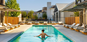 Woman in a pool with arms outstretched, surrounded by lounge chairs and a white building.