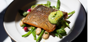 Pan-seared fish on a white plate with greens, beans, radishes, and green sauce.