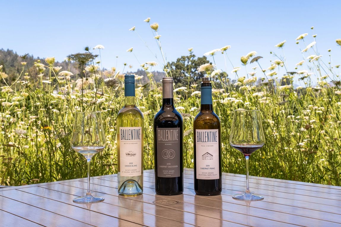 Three wine bottles on a table with two empty wine glasses, set in a field of wildflowers.