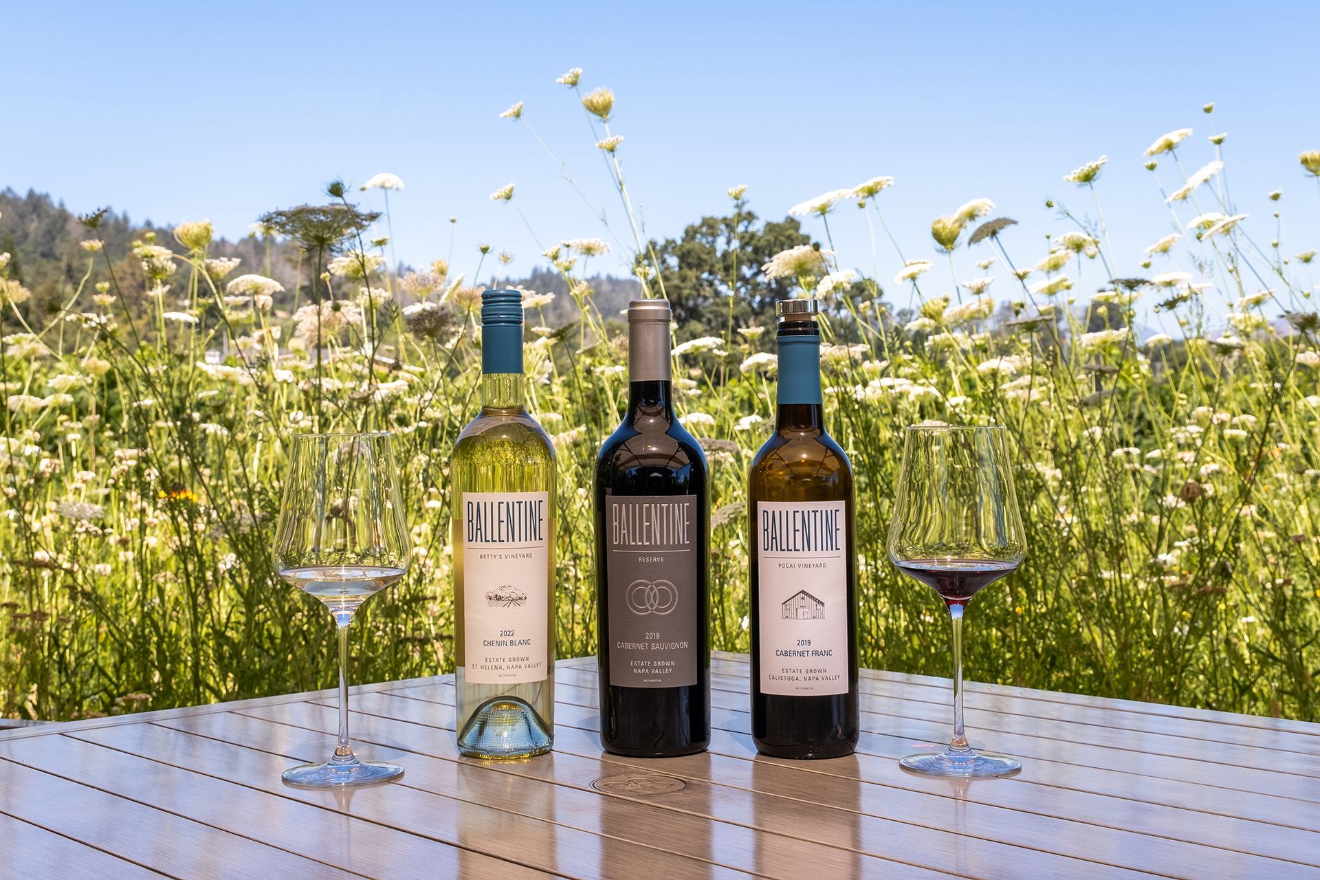 Three wine bottles on a table with two empty wine glasses, set in a field of wildflowers.