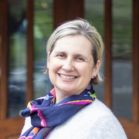Woman with blonde hair and a colorful scarf smiles in front of a wooden door.
