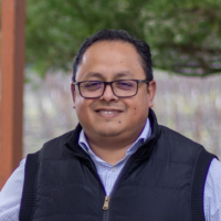 Man wearing glasses and vest, smiling outdoors.