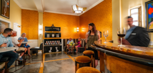 People at a bar, with wine glasses and stools. The setting has yellow walls.