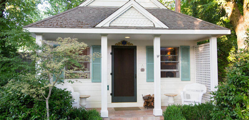 White cottage with gray roof, green shutters, and a dark door, surrounded by lush greenery.