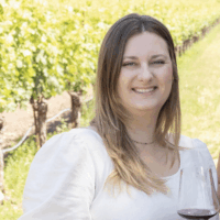 Woman smiling in vineyard, holding wine glass. Sunlight, white top.