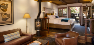Cozy hotel room with a fireplace, bed, and outdoor balcony. Brown leather furniture, warm lighting, and a neutral color palette.