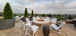 Patio with fire pit, white chairs, potted trees, and cloudy sky.