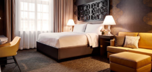 A hotel room with a bed, desk, sofa, and window with curtains. Warm lighting and neutral colors.