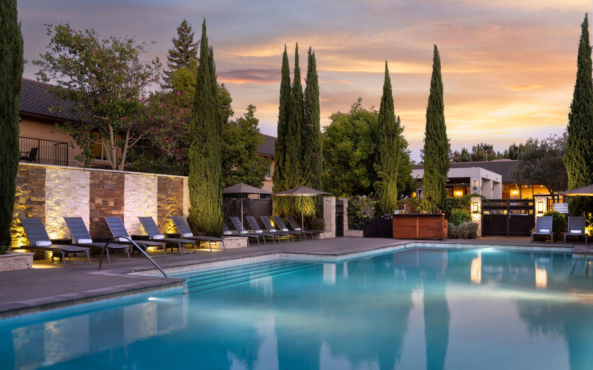 Poolside lounge chairs, cypress trees, and a pool at sunset.