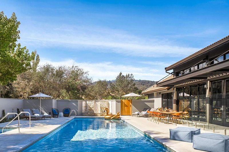 Pool with lounge chairs, dining table, and building under a bright blue sky.
