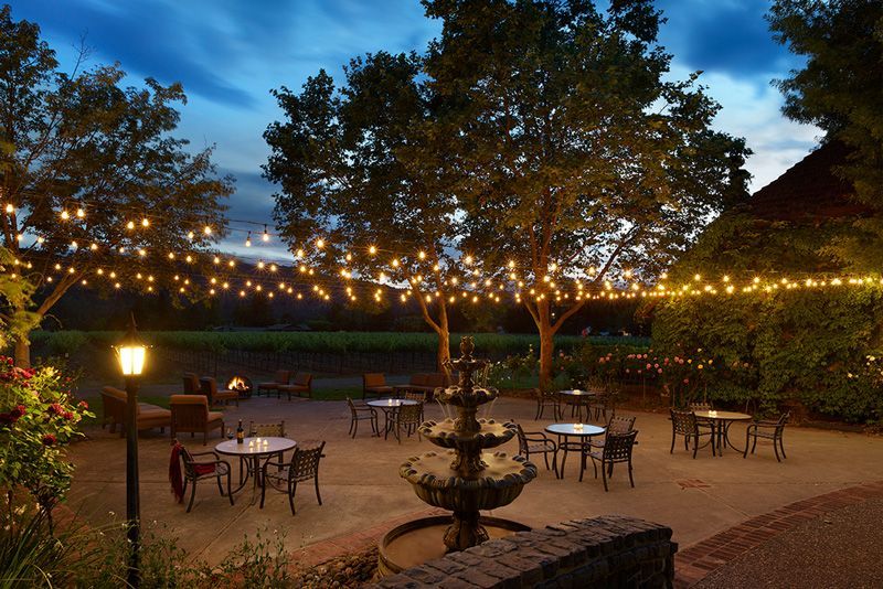 Outdoor patio at dusk, string lights, tables, chairs, fountain, and trees.